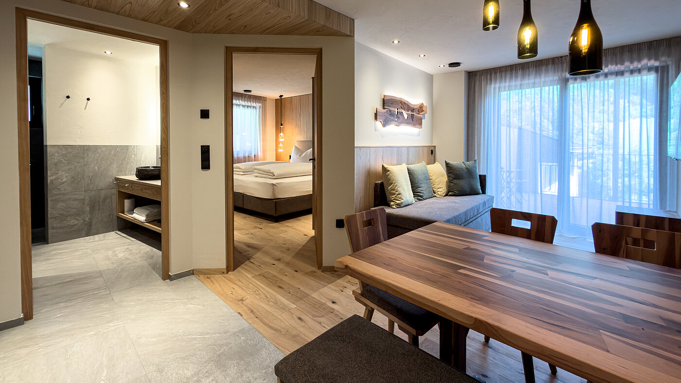 Offener Wohnbereich im Bio-Wein-Apartment – Weingut Santer Blick vom offenen Essbereich mit Massivholztisch auf das Sofa, das angrenzende Schlafzimmer und das Badezimmer im Weingut Santer.