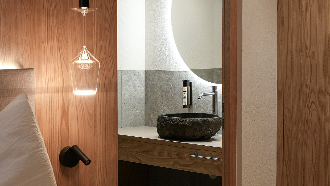 Modernes Badezimmer im Wein-Apartment – Bio-Weingut Santer Modernes Badezimmer mit Natursteinwaschbecken und beleuchtetem Spiegel, gesehen vom Schlafbereich aus im Weingut Santer.
