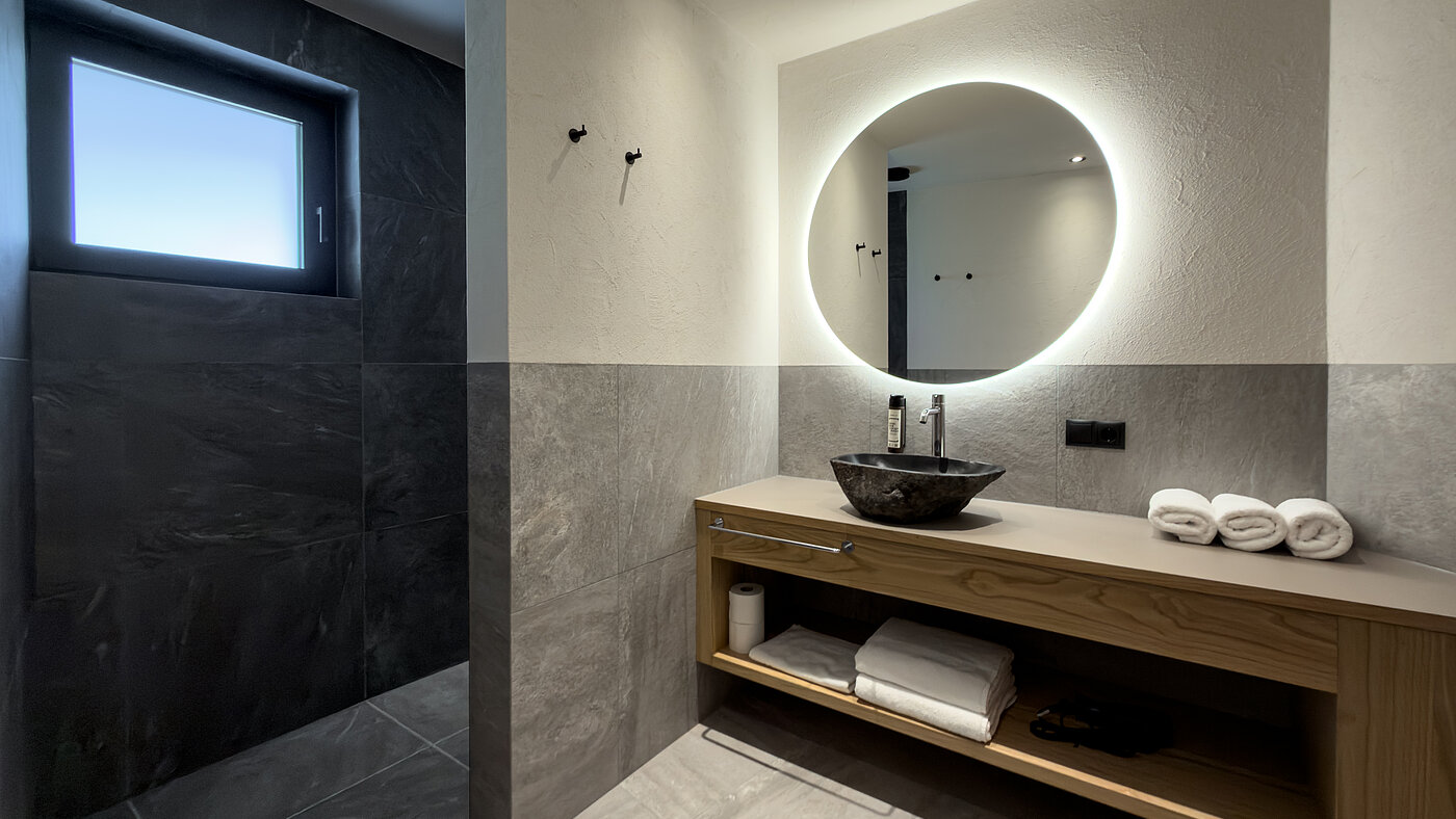 Badezimmer mit Tageslicht im Wein-Apartment – Weingut Santer Modernes Badezimmer im Weingut Santer mit rundem LED-Spiegel, Fenster für Tageslicht, Steinwaschbecken und offener Dusche.