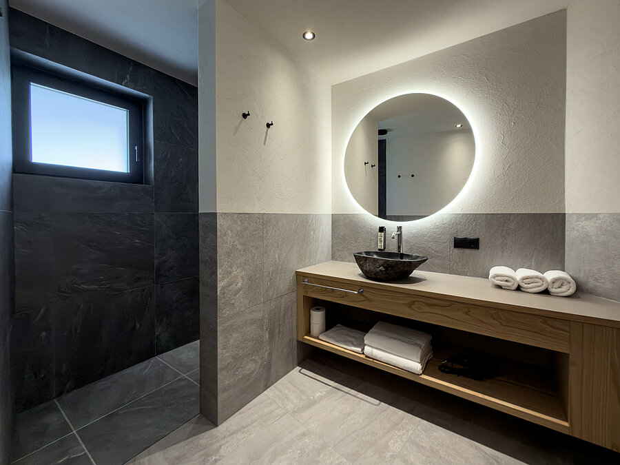 Badezimmer mit Tageslicht im Wein-Apartment – Weingut Santer Modernes Badezimmer im Weingut Santer mit rundem LED-Spiegel, Fenster für Tageslicht, Steinwaschbecken und offener Dusche.