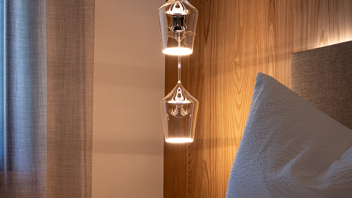 Kreative Weinglas-Leuchte im Schlafzimmer – Weingut Santer Detailansicht einer Hängelampe aus Weingläsern neben einem Bett vor einer gemaserten Holzwand im Weingut Santer.