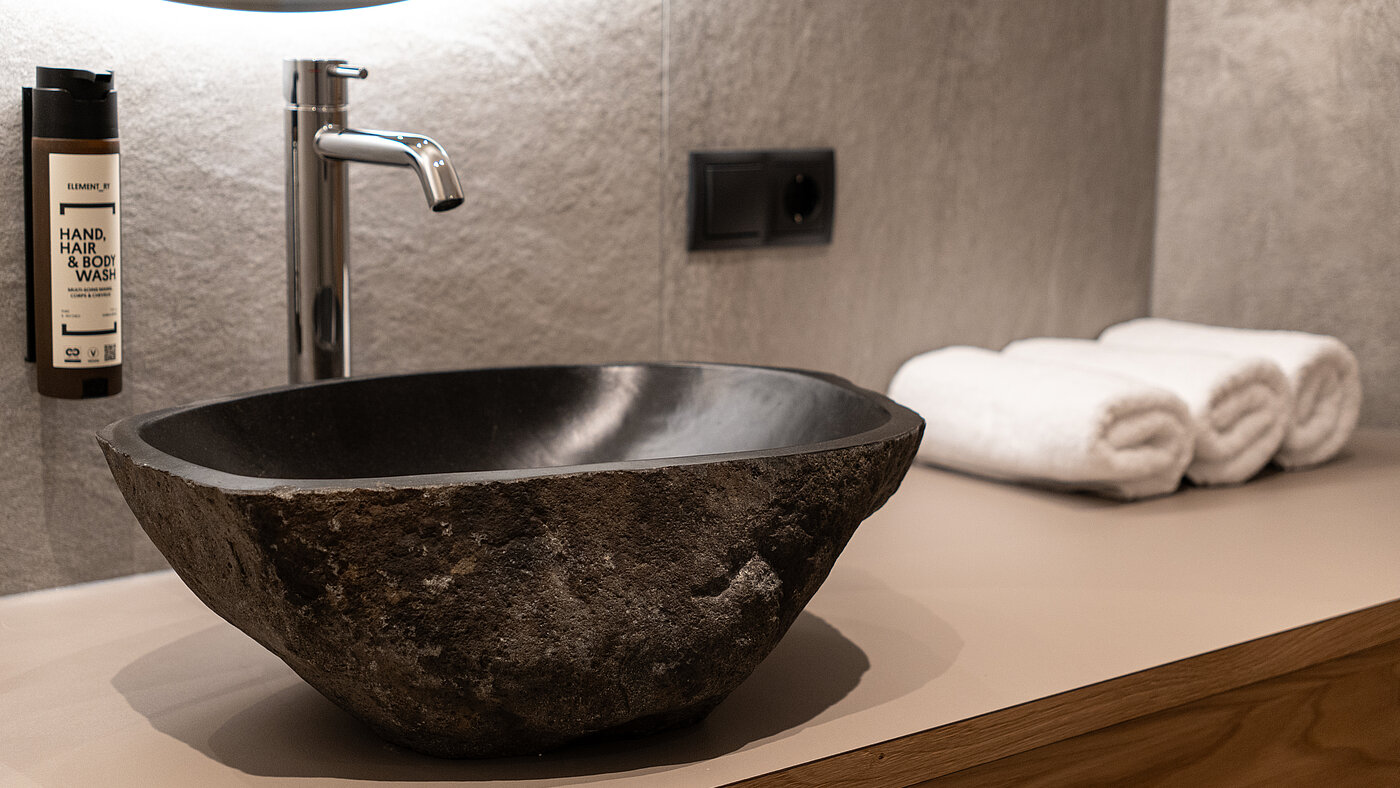 Naturstein-Waschbecken im Wein-Apartment – Weingut Santer Nahaufnahme eines rustikalen, dunklen Naturstein-Waschbeckens auf einem Holzwaschtisch mit weißen Handtüchern im Weingut Santer.