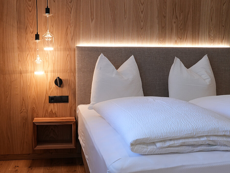 Schlafzimmer aus Naturholz im Wein-Apartment – Bio-Weingut Santer Modernes Doppelbett vor einer Wand aus Naturholz mit hängenden Designerleuchten aus Weingläsern im Weingut Santer.IT