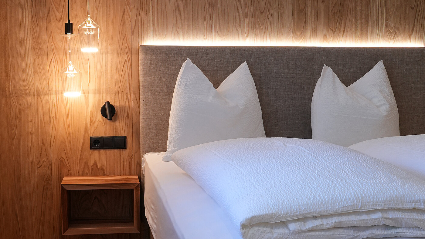 Schlafzimmer aus Naturholz im Wein-Apartment – Bio-Weingut Santer Modernes Doppelbett vor einer Wand aus Naturholz mit hängenden Designerleuchten aus Weingläsern im Weingut Santer.IT