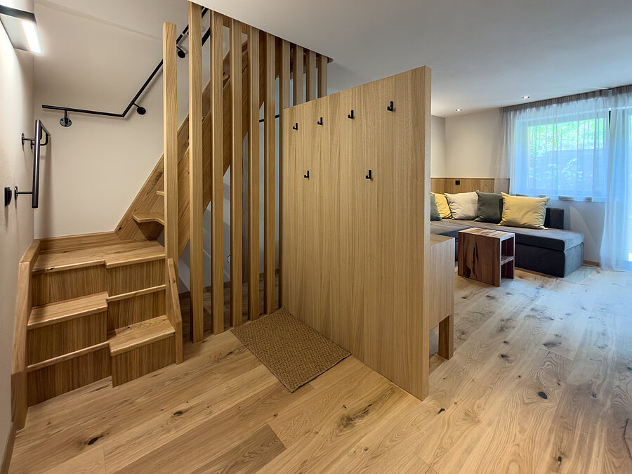 Eingangsbereich mit Holztreppe im Wein-Apartment – Weingut Santer Eingangsbereich einer Ferienwohnung im Weingut Santer mit moderner Massivholztreppe, hölzernem Raumteiler und Blick auf das Sofa.