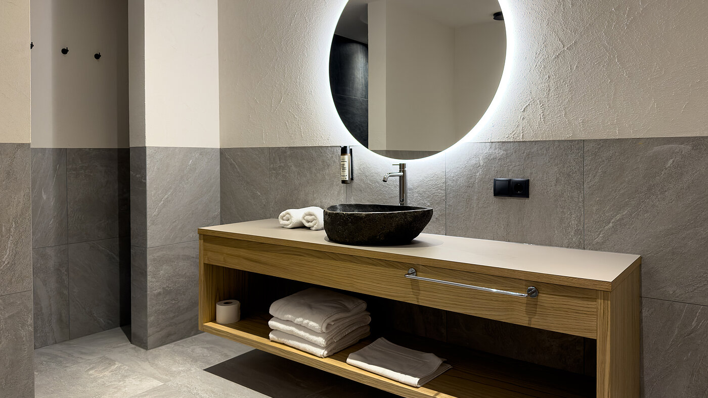 Design-Badezimmer im Wein-Apartment – Bio-Weingut Santer Modernes Badezimmer im Weingut Santer mit rundem LED-Spiegel, strukturierter Mosaikwand, Steinwaschbecken und Holzwaschtisch.