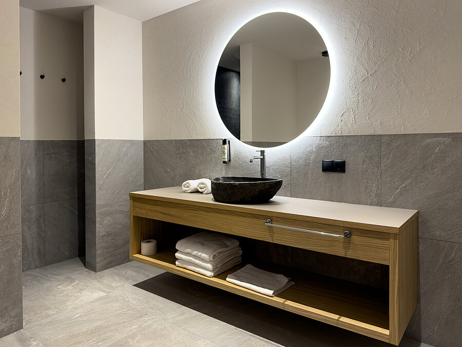 Design-Badezimmer im Wein-Apartment – Bio-Weingut Santer Modernes Badezimmer im Weingut Santer mit rundem LED-Spiegel, strukturierter Mosaikwand, Steinwaschbecken und Holzwaschtisch.