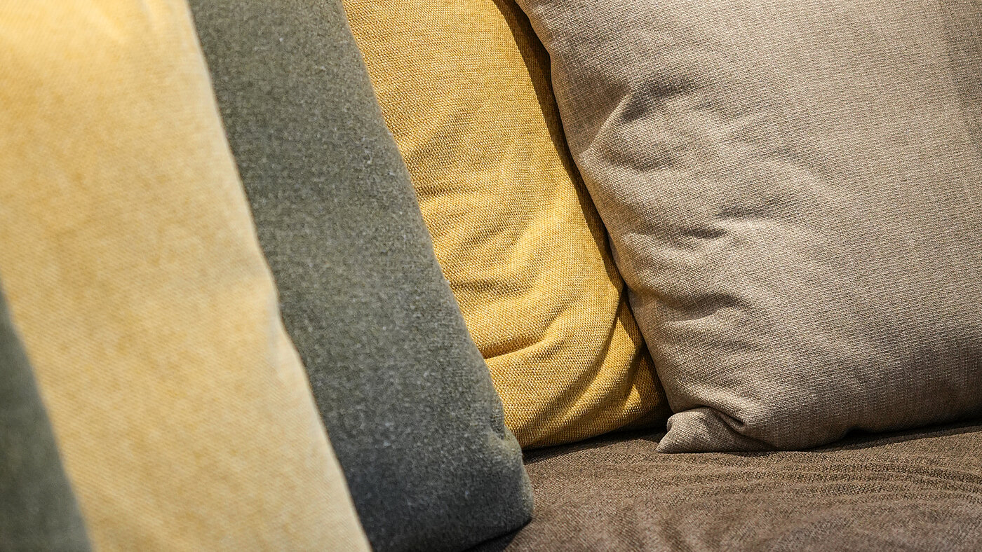 Gemütliche Sofakissen im Wein-Apartment – Weingut Santer Detailaufnahme von gemütlichen, farbigen Dekokissen in Naturtönen auf einem Sofa vor einer Holzvertäfelung im Weingut Santer.