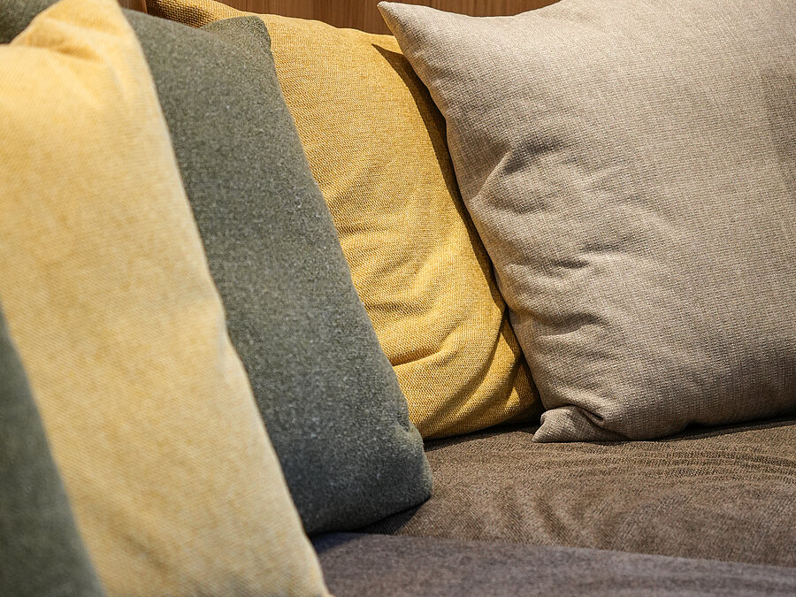 Gemütliche Sofakissen im Wein-Apartment – Weingut Santer Detailaufnahme von gemütlichen, farbigen Dekokissen in Naturtönen auf einem Sofa vor einer Holzvertäfelung im Weingut Santer.
